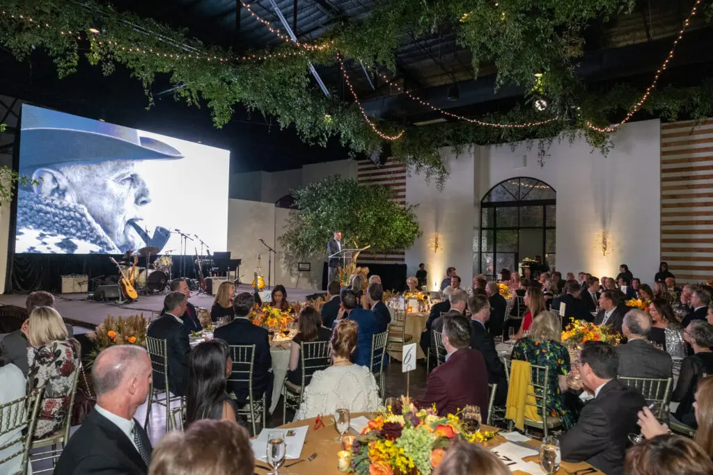 Ucross president Bill Belcher addresses the gathering at The Revaire where an image of founder Raymond Plank filled the screen. (Photo by Jenny Antill)