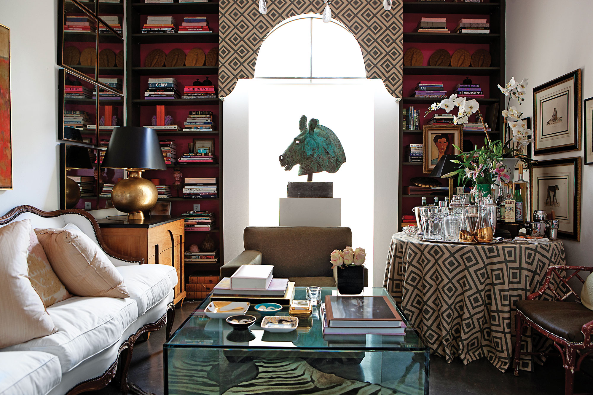 The library/den is centered around a Roman bronze horse head from Biff Agee, which sits atop a custom wooden stand. Valence is covered in David Hicks La Florentina from Lee Jofa\Kravet, flanked by a pair of bookshelves painted olive with hot-pink interior. A David Hicks-inspired drinks table holds a large silver tray, Baccarat ice bucket, collection of vintage crystal shakers and decanters. Antique hand colored prints were gifts from Cathy’s mother. Faux-bamboo chair covered in olive-colored cowhide. Zebra rug from Custom Creations is beneath a custom glass cube coffee table. An 18th-century French Louis XIV sofa from James Powell is flanked by Donghia chests and Robert Kuo hammered-gold, washed-bronze lamps from Ellouise Abbott.
