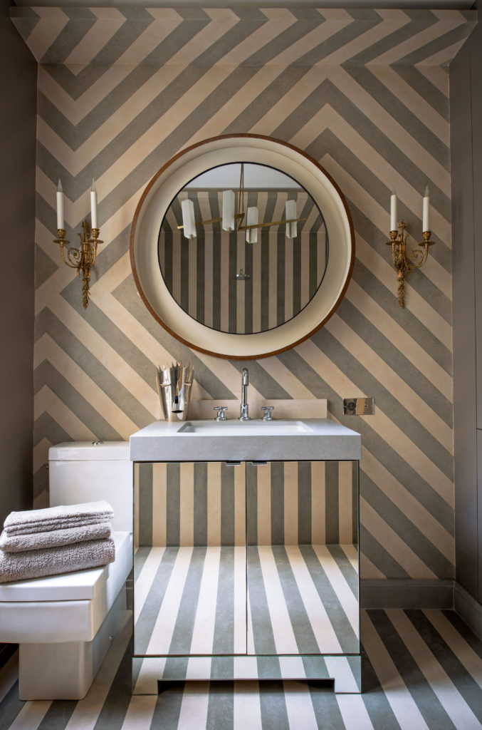 In a guest bath on rue de Lille, French limestone and Portuguese Cascais stone cut into stripes in the style of French artist Daniel Buren. 1950s Danish backlit mirror, bronze Louis XVI wall lights.
