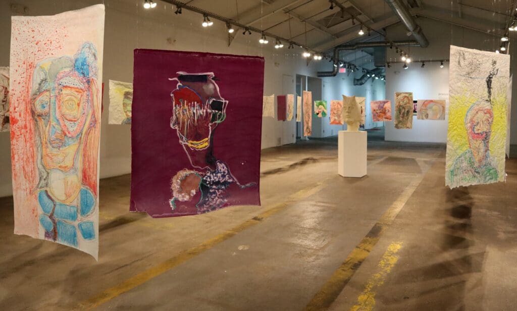 Justin Earl Grant subverts the traditional art installation by having his colorful portraits of distorted face hung and hovering around the viewer. 