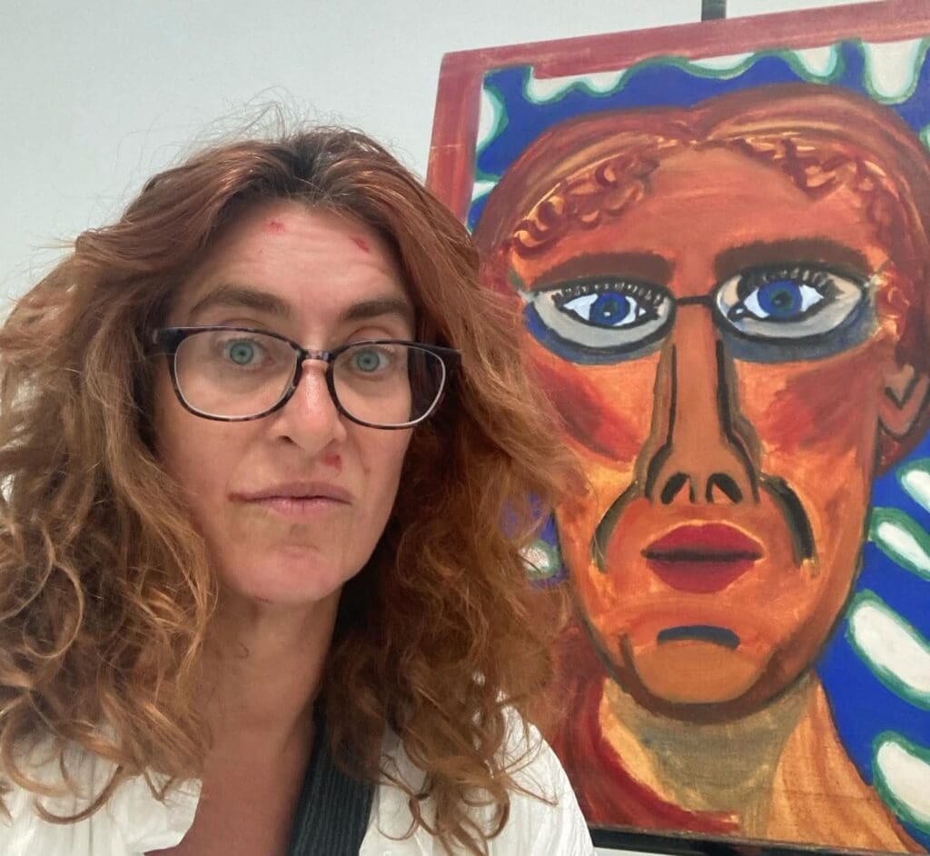 Artist Paula Liftschitz standing next to a self-portrait she did in her first painting class in college at the University of Pennsylvania. 