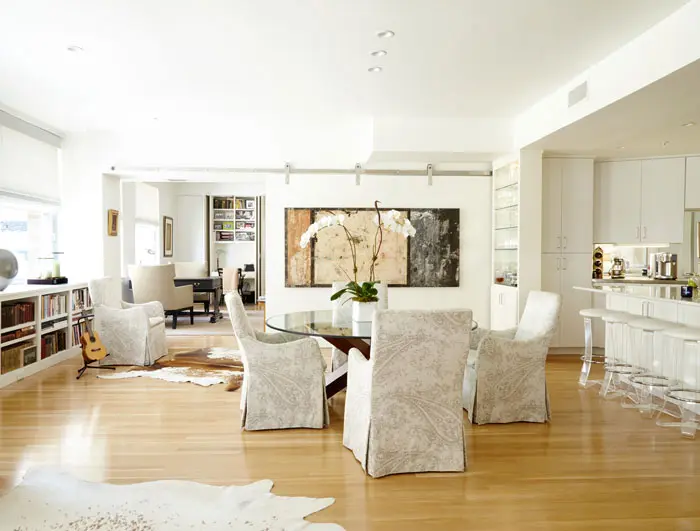 In the dining area, a dining table from Scott + Cooner is attended by chairs upholstered in Peacock Alley linen fabric. The vintage Lucite stools were found at an antique store in Dallas and reupholstered in white ostrich. A custom sliding wall acts as a room divider and backdrop for a large painting by Santa Fe artist Marcia Myers. The floors are white pine.