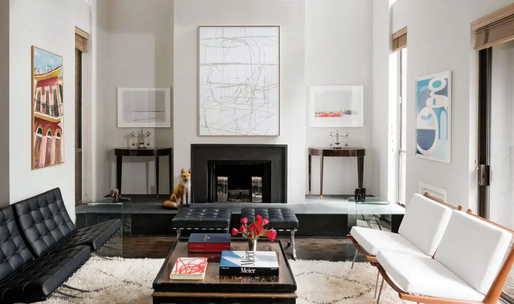 The home’s shared living and dining room acts as its formal entrance. Two tufted black-leather Mies van der Rohe Barcelona chaises and matching stools pair with a lacquered coffee table atop one of many Moroccan rugs Gordon acquired while visiting that country for a friend’s wedding. Pair of white Boomerang lounge chairs by Peter Hvidt and Orla Mølgaard- Nielsen. Artwork, including pieces by Aaron Parazette and Anton Ginzburg, gives the buoyant environment an intriguing air. Alcoves on either side of the fireplace frame Deco consoles and sterling Edwardian candelabra.