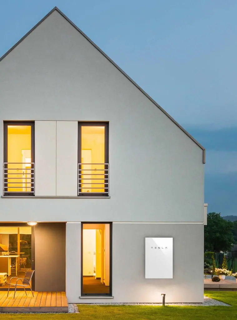 A Tesla Powerwall 2 home battery, shown on an exterior house wall, stores energy from solar panels.