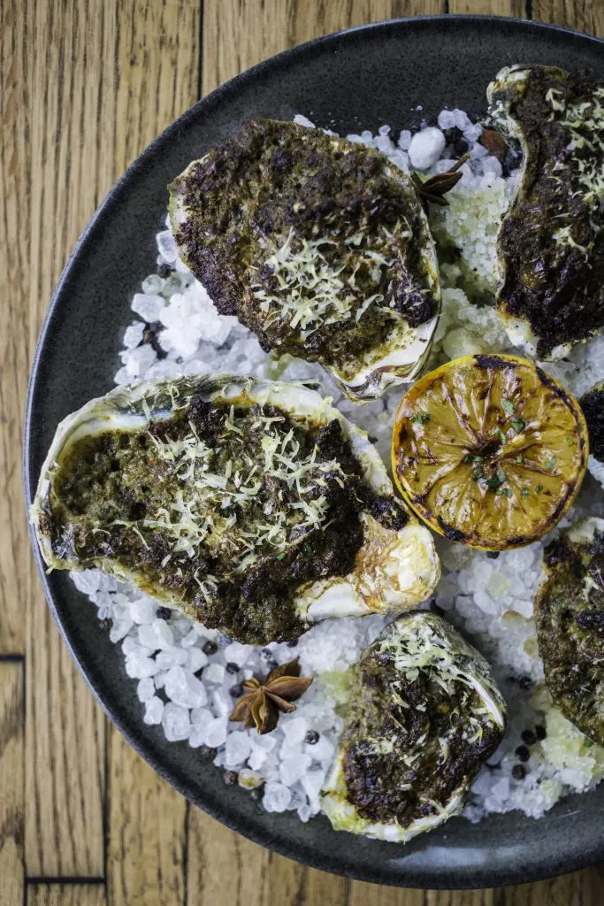Baked Oysters made with tomato, onion, pesto, parmesan, bacon, and aromatic rock salt. 