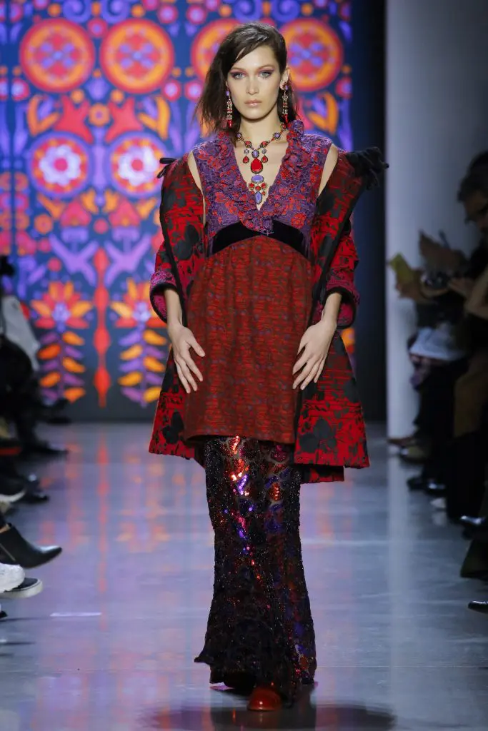 Model Bella Hadid in Anna Suit rouge multi-gradient floral jacquard coat, festive sequin pant, and multi flowers from a fan lace and jacquard dress. 
