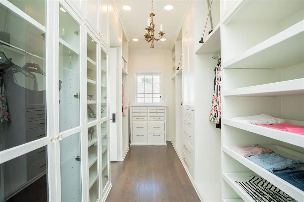 The spacious hers closet has white oak floors, grass front cabinets, slide-out shelves, a chest of drawer with a white marble top and more. 