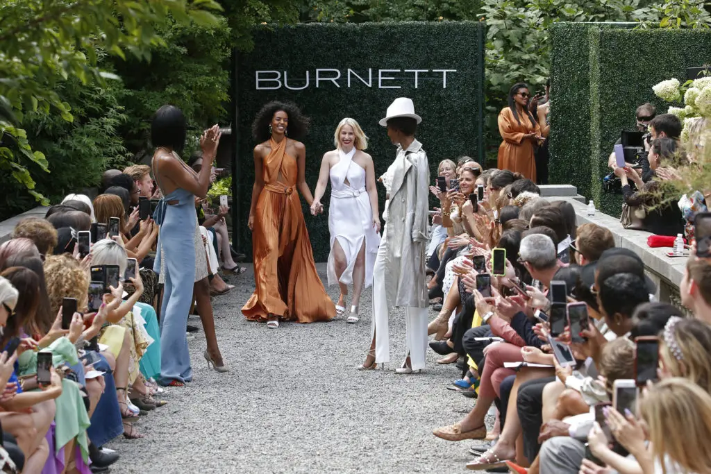 Emily Burnett, middle in white pantsuit, takes her runway bow with model Grace Mahary, in hazelnut charmeuse pleated halter gown. (Photo courtesy of Burnett New York)