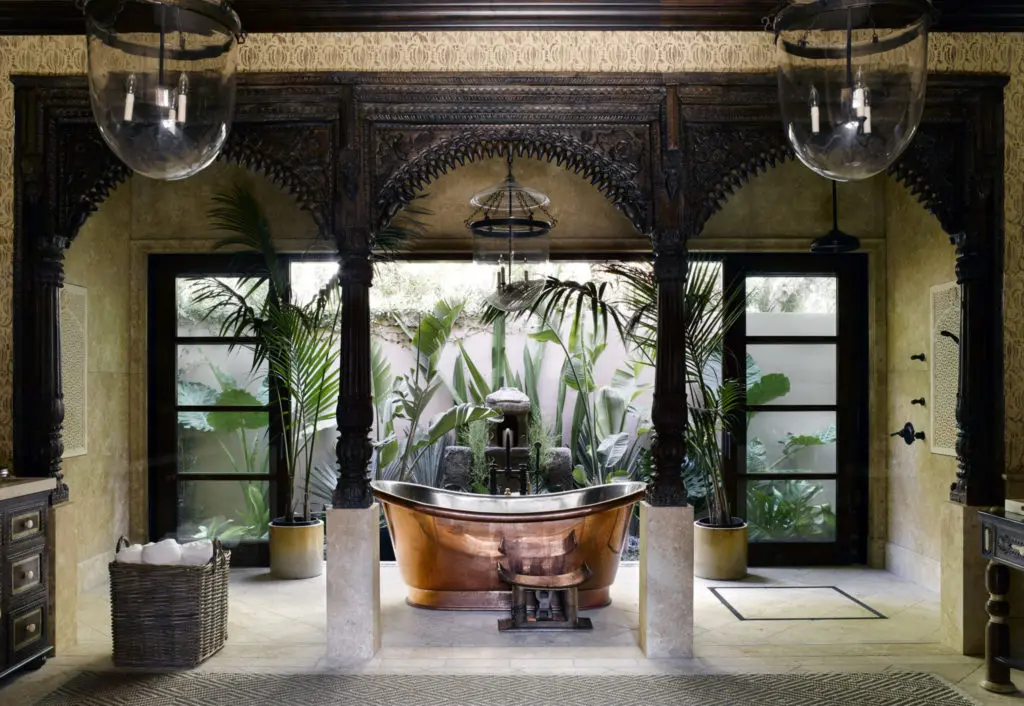 In a Mediterranean-style home in Malibu, the master bathroom was created from the facade of a Rajasthani palace. The copper bathtub is from Waterworks, and the wallpaper is by Rose Tarlow. The giant glass Hundi lanterns were custom blown for the space. An African headrest is perfect for placing a drink or laying a towel by the side of the bath.