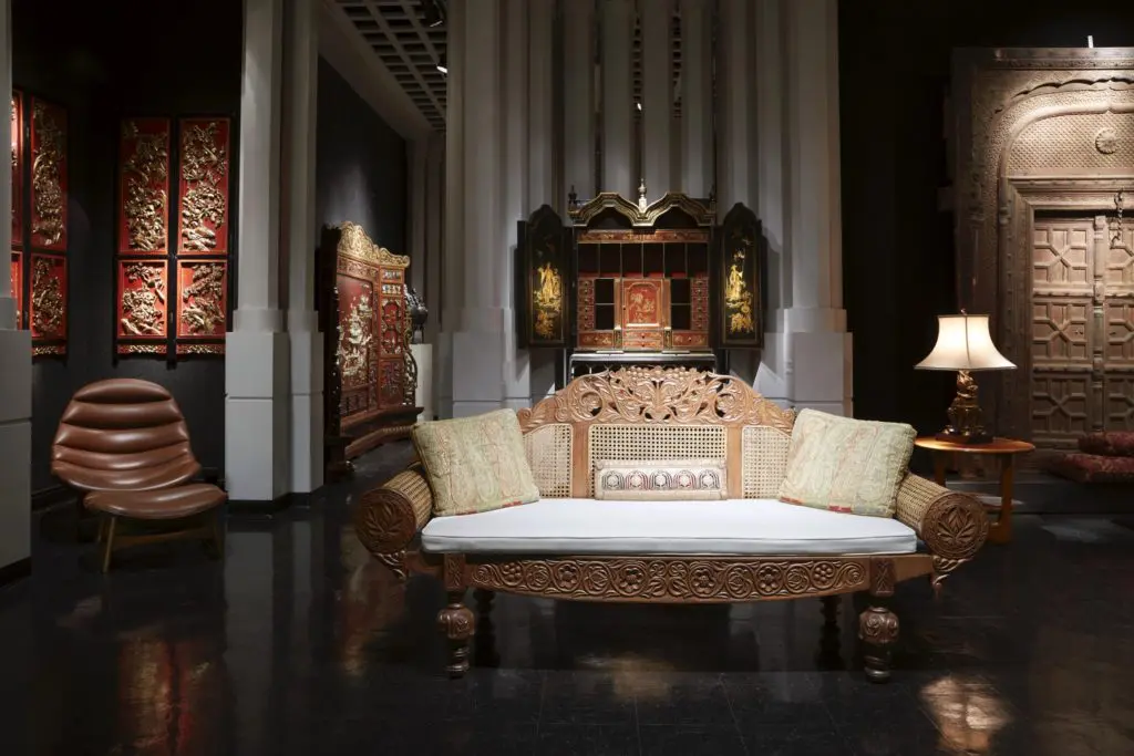 A British Colonial satinwood and cane settee, ornately carved with scrolling foliage, vines, and flowers. An English chinoiserie secretary from the early 20th century has gilt painted scenes of Chinese figures, pagodas, and gardens. (Photo by Pär Bengtsson)