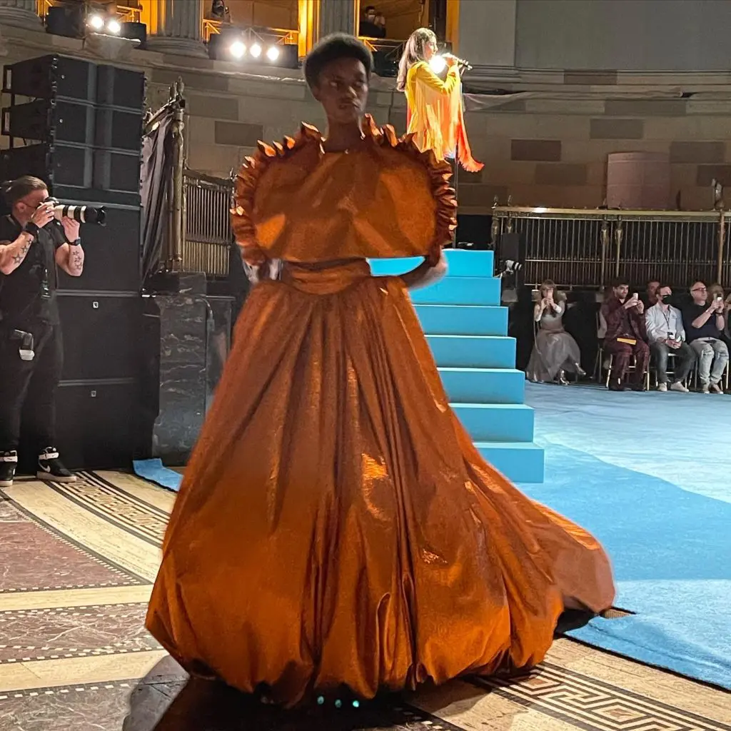 Christian Siriano sculpted gold gown. Welsh singer Marina Diamandis, shown in background, performed during the runway show. (Photo by Clifford Pugh)