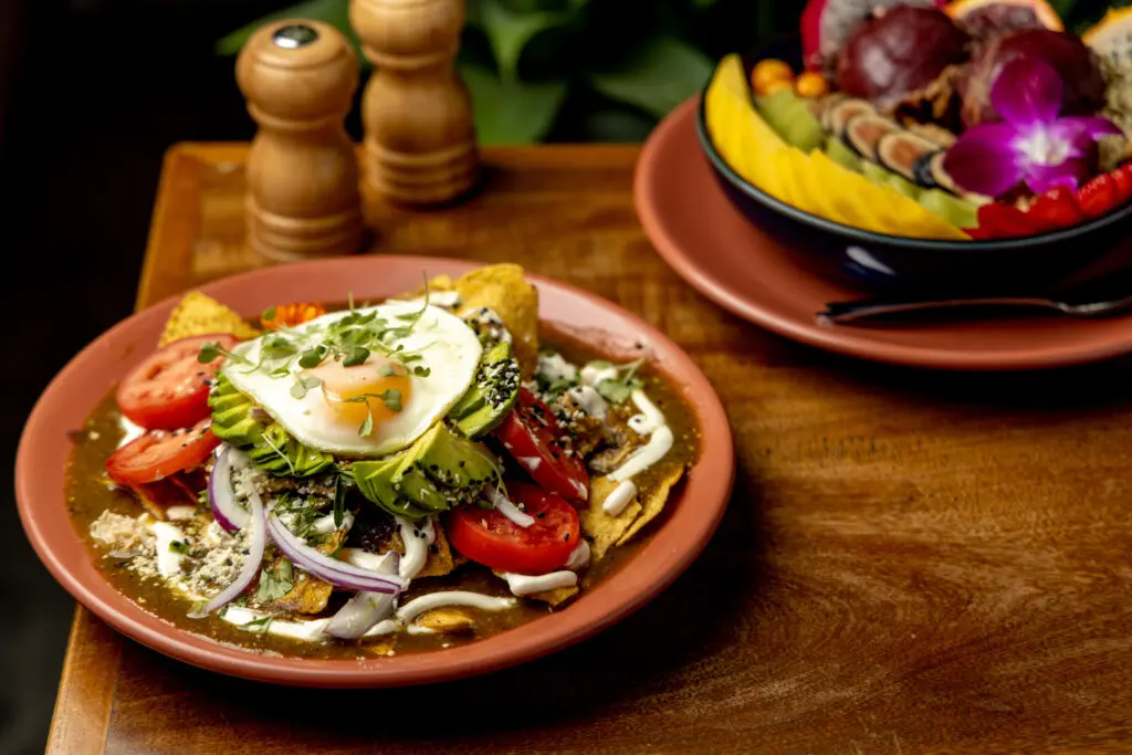 The red (rojos) chilaquiles at Ojo de Aqua are served with chicken ($22) baked and layered with tortilla quarters, spicy rojo sauce, beans, shredded cotija cheese, tomato, onion, and topped with avocado slices and sesame seeds. (Photo by Jennifer Hasbún)