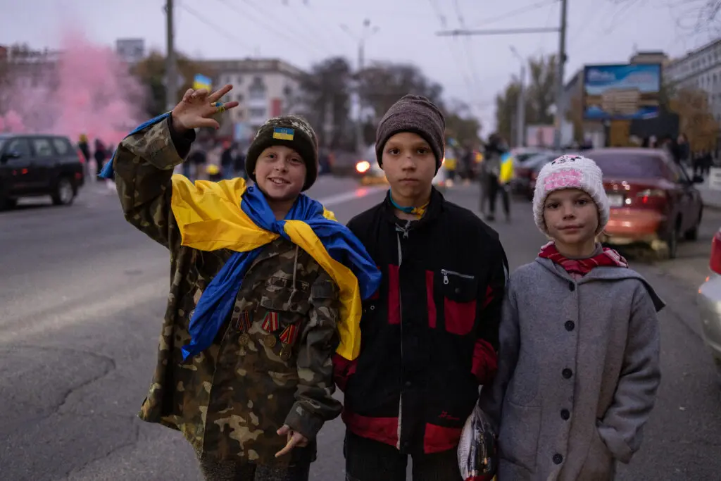 Children with a Ukrainian flag and uniforms flash the victory sign on the central square of Kherson during celebrations of the city’s liberation by Ukrainian army. November 12, 2022.(Photo by Andriy Dubchak. Courtesy Ukrainian Association of Professional Photographers) 
