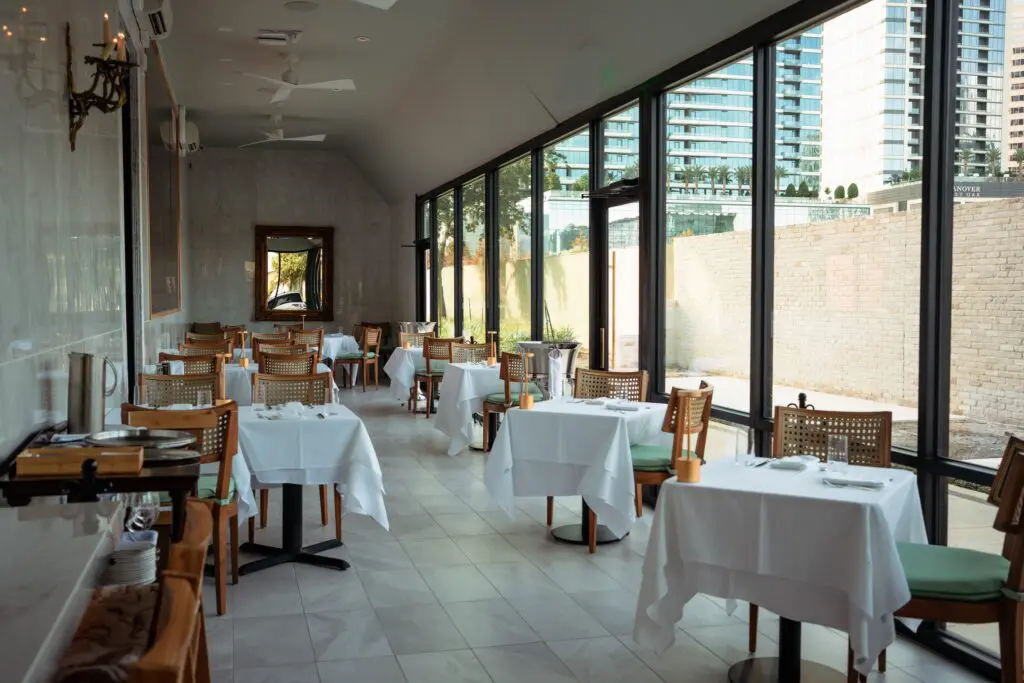 Sunny afternoons invite dining in the glass-walled seating area at Tavola, adjacent to the main dining room replete with vibrant green striped covered cane back chairs.