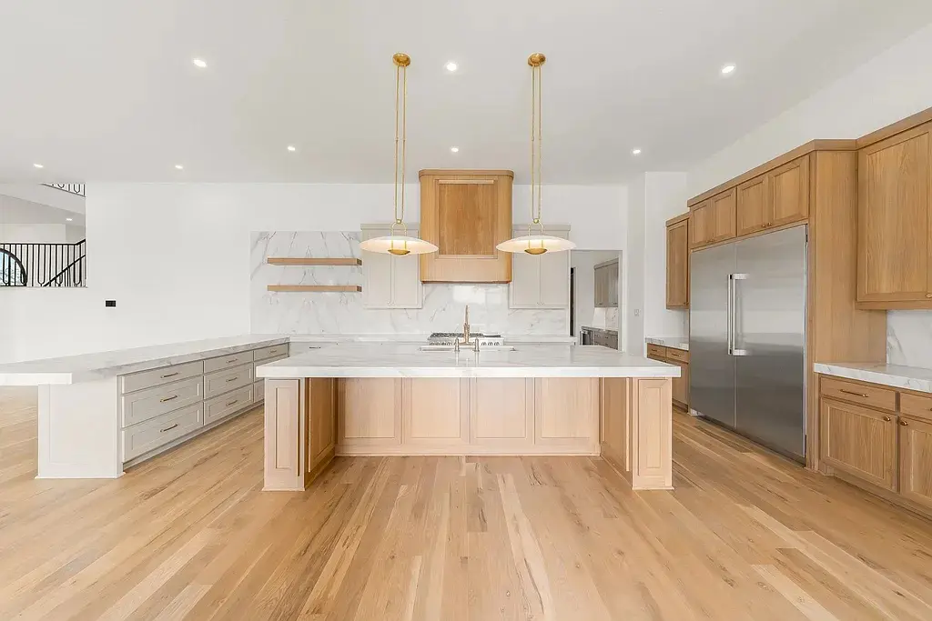 The kitchen features an oversized waterfall island, brass pendant lighting, marble-style surfaces, and extensive light wood cabinetry. (Courtesy)