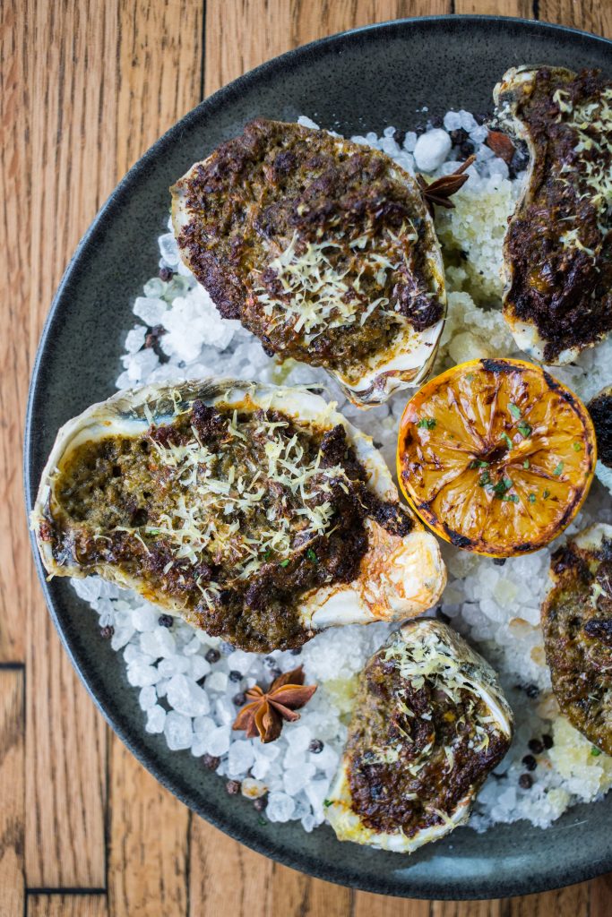 Baked Oysters made with tomato, onion, pesto, parmesan, bacon, and aromatic rock salt.