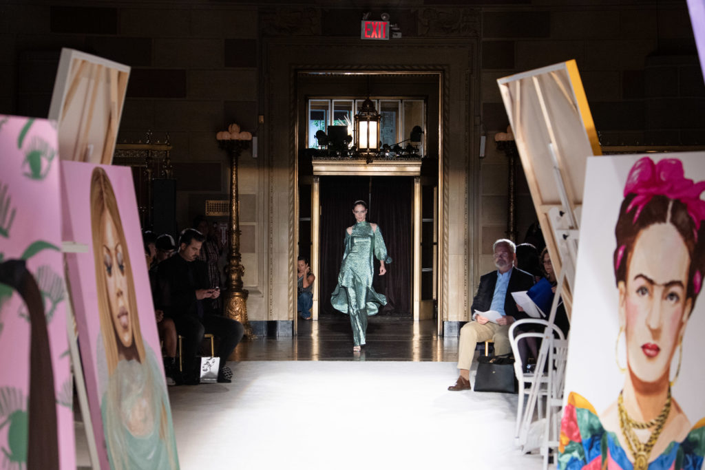 Model Coco Rocha opens the Christian Siriano runway show in a seafoam shimmer cut-out asymmetrical dress  and flared trousers at New York Fashion Week amid a slew of paintings by Ashley Longhore. (Photo by Luca Zanoni)