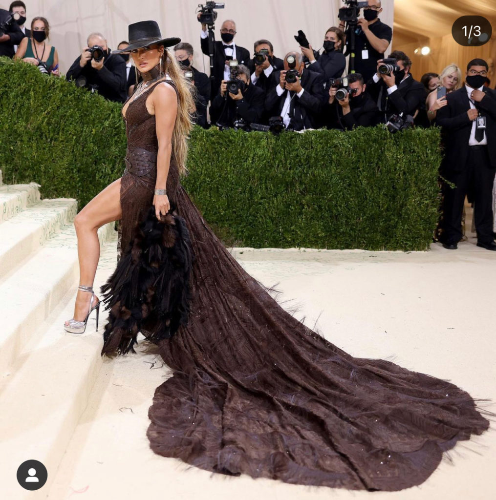Jennifer Lopez wears Ralph Lauren gown with a plunging neckline, feathered trim, thigh-high slit, and cowgirl hat. She paired the look with metallic silver strappy stilettos from her DSW line. (instagram: jlo)