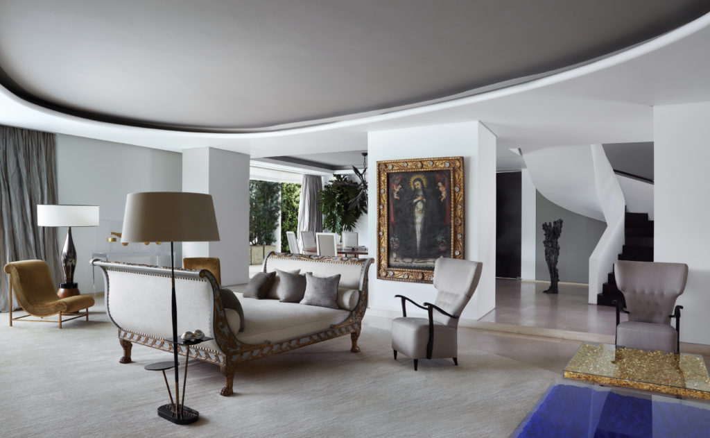  In Cali, Columbia, accessories designer Nancy Gonzales’s living room, designed by Jean-Louis Deniot, is both graceful and surprising, with a seventeenth-century Madonna, and iconic Yves Klein blue and gold-leaf tables. Pair of Rita armchairs by Jean-Louis Deniot for Collection Pierre upholstered in cotton velvet by Romo. Mid-century French brass and black opaline asymmetrical floor lamp with tabletops. Empire daybed in grey patina and gilt accent in Romo linen, and custom-designed JLD rug by Diurne.