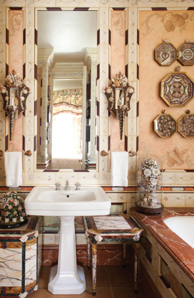 The Josephine guest bathroom is embellished with scrolls and flourishes of Victorian penwork and delicately rendered faux-ivory and ebony framing. The room offers delight to the eye with a pair of brilliant pieta dura tables, along with verre églomisé panels of coral and seaweed. On the bath surround is one of three glass-domed shellwork floral bouquets, mid-19th century, with elaborate flowers worked in seashells and wire supports. The Victorian-era octagonal hinged boxes are framed sailors' valentines, a West Indies tradition. The pair of French cartouche-shaped shellwork sconces is French. (Photography by Lisa Romerein)