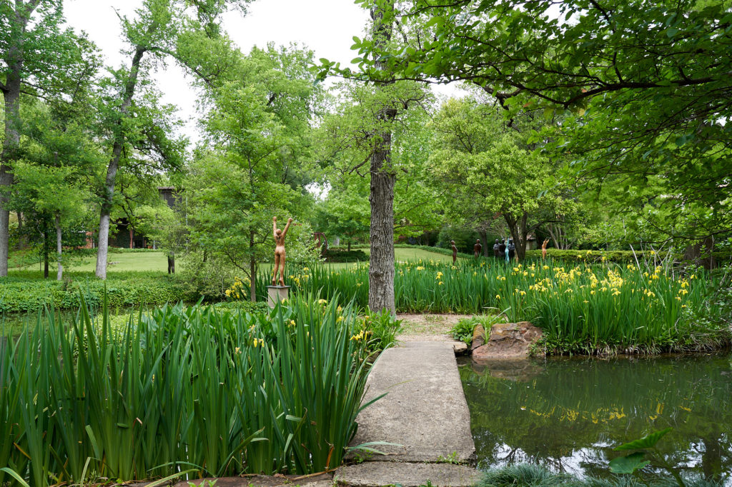 An island in the pond holds a sculpture by Deborah Ballard. The concrete bridge was designed by Donald Vogel. Yellow Louisianna irises growing in the water. (Photo by Pär Bengtsson / art direction by Michelle Aviña)

