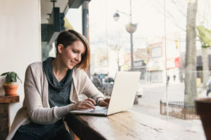 Female customer sitting in coffee shop window seat typing on laptop