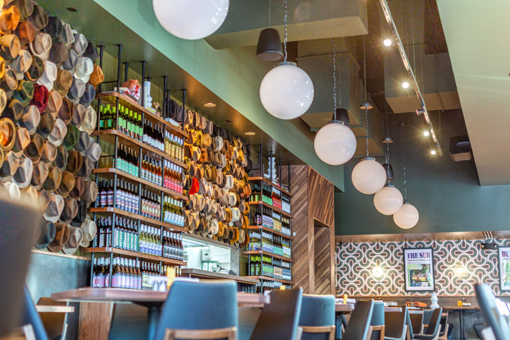 The art wall at Postino Woodlands os decorated with hundreds of colorful, authentic 1950s and 1960s era fedoras sourced from a vintage picker in Round Top as a nod to Howard Hughes’ love of aviation. (Photo by Jenn Duncan)
