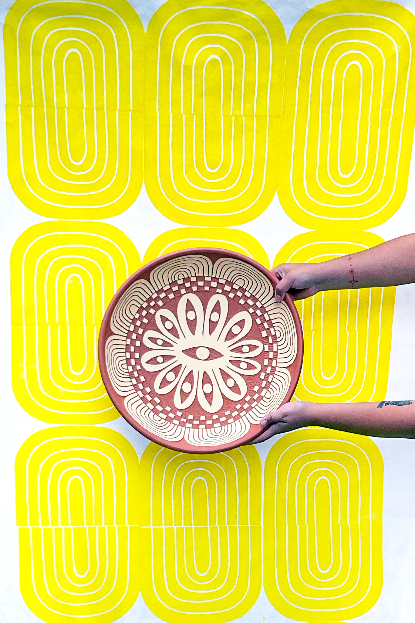 Gabo Martinez holding “Twelve Eyes Platter” (inside view), 2023. Background: “Yellow Bloom Print,” 2021. (Photo courtesy the artist)