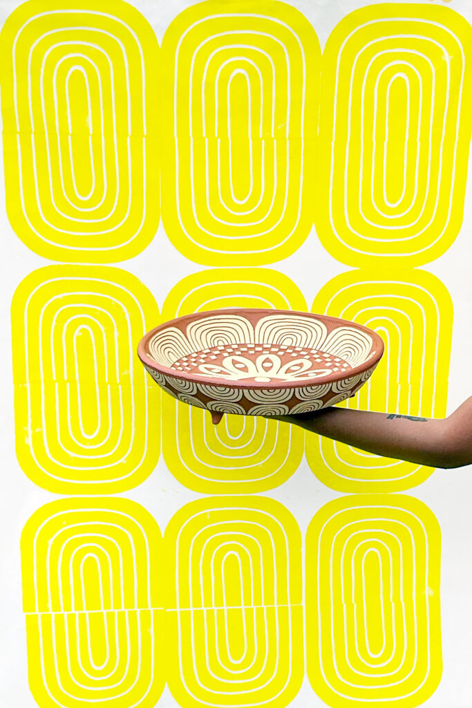 Gabo Martinez holding “Twelve Eyes Platter," 2023. Background: “Yellow Bloom Print,” 2021. On view at Houston Center for Contemporary Craft (Photo courtesy of the artist)
