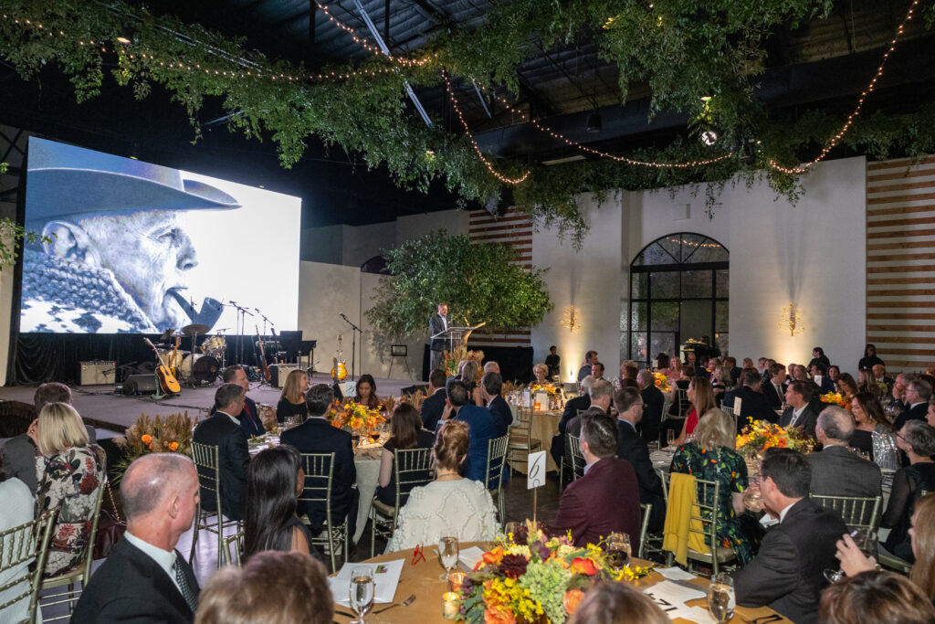 Ucross president Bill Belcher addresses the gathering at The Revaire where an image of founder Raymond Plank filled the screen. (Photo by Jenny Antill)
