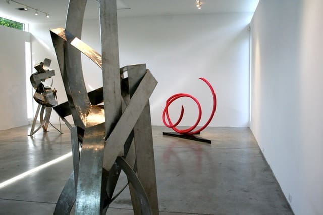 An installation view of the "Heavy Metal" exhibition in 2015, which featured sculptures by artists Mac Whitney and John Clement at Gallery Sonja Roesch, Houston. The two metallic sculptures on the left were made by Whitney. Clement created the red curvilinear sculpture on the right. (Courtesy Gallery Sonja Roesch, Houston)