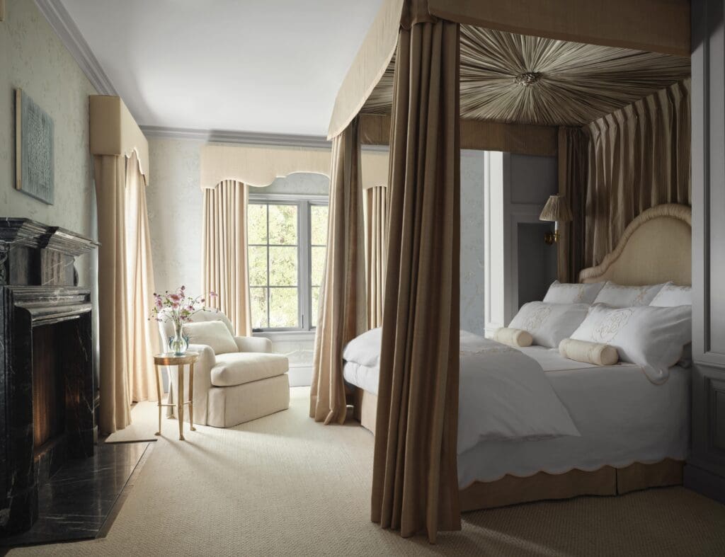 The main bedroom is anchored by an antique marble fireplace and custom canopy bed, draped in Colefax & Fowler slubbed silk. De Gournay wallpaper. (Photo by Douglas Friedman)