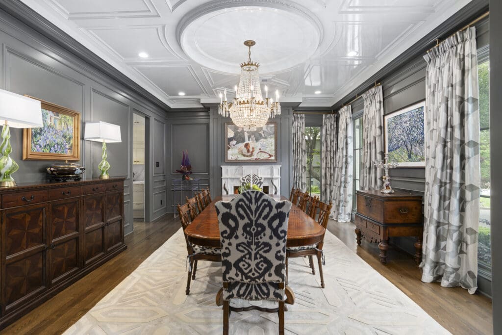 A coffered ceiling and crystal chandelier set the tone in the formal dining room, where rich paneling, layered drapery, and classic furnishings create timeless appeal. (Freeze Media)