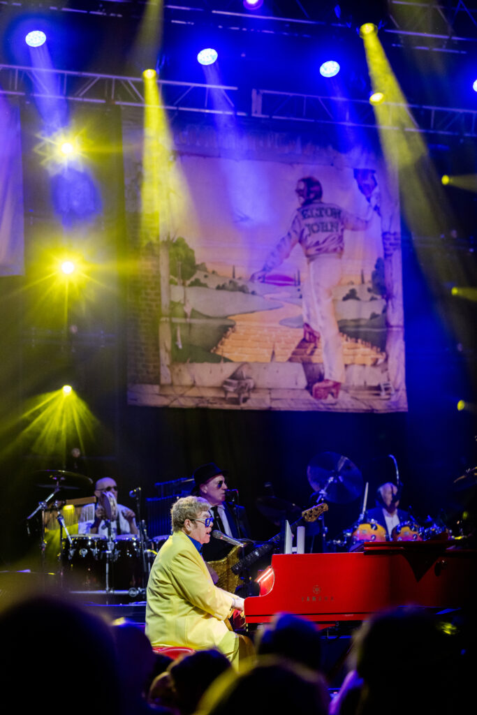 Without any announcement over the speakers, a massive curtain fell to the stage floor, dramatically revealing Elton John dressed in an electric yellow jacket behind the piano. (Photo by Anastasiia Photography)